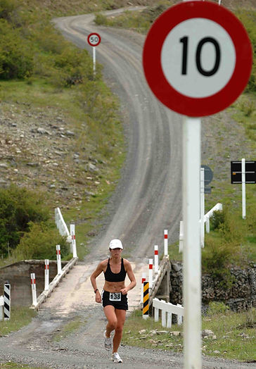 A solo woman runner runs toward the camera up a gravel road. She has just crossed a single lane wooden bridge. There is a prominant 